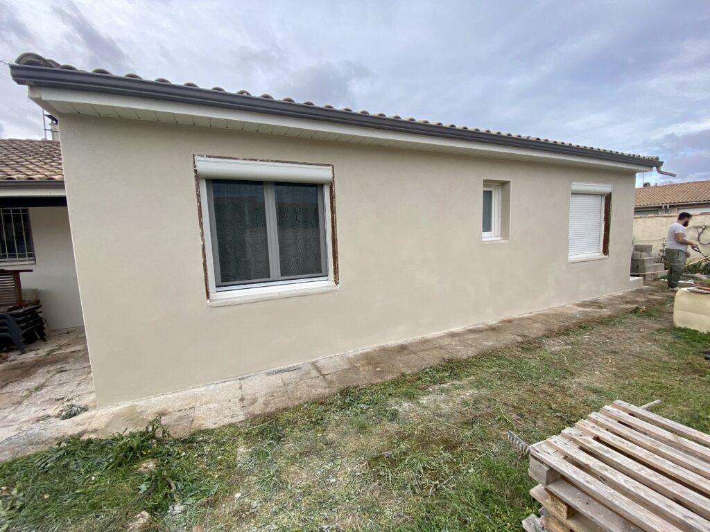 Maison de plain-pied avec façade fraîchement rénovée en enduit clair, fenêtres en cours de finition, et artisan façadier au travail sur le côté droit.