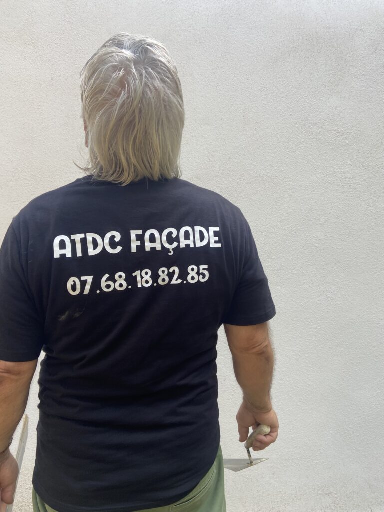 Artisan façadier vu de dos portant un t-shirt noir avec l'inscription "ATDC Façade" et un numéro de téléphone, tenant une taloche devant un mur lisse en cours de rénovation.